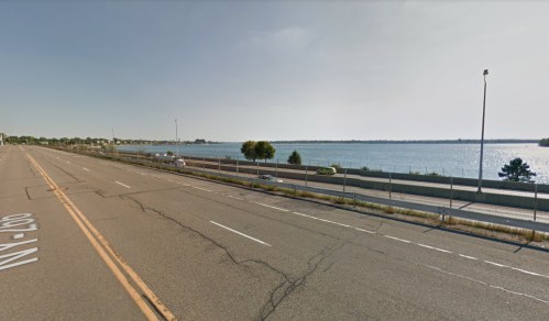 A view of a road alongside a body of water, with trees and a clear sky in the background.