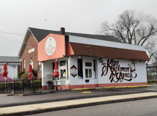 A brick building with a sign reading "Solomon Gyros," featuring outdoor seating and a cloudy sky.