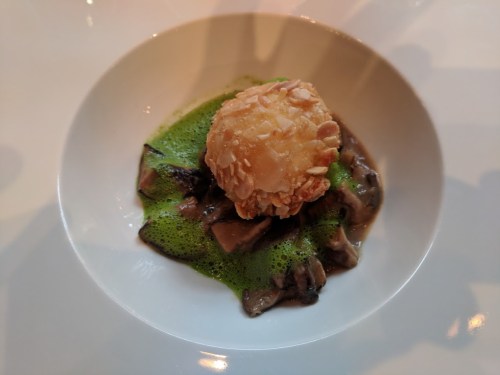 A gourmet dish featuring a crispy ball atop a bed of mushrooms and vibrant green sauce.