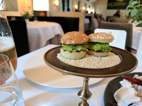 Two mini burgers with avocado on a decorative stand, served in a restaurant setting with a glass of beer nearby.