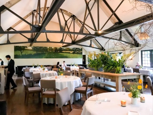 A stylish restaurant interior with high ceilings, wooden beams, and tables adorned with flowers and candles.