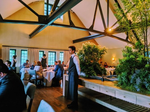 A stylish restaurant interior with diners, a waiter, and greenery decor under a high ceiling with wooden beams.