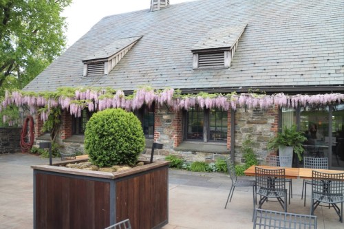 A charming stone building with a slate roof, adorned with purple wisteria and surrounded by outdoor seating.