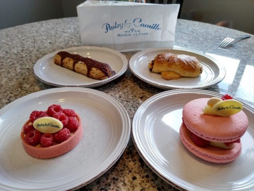 Four pastries on white plates: a raspberry tart, a chocolate croissant, a scone, and a pink macaron, with a pastry bag in the background.