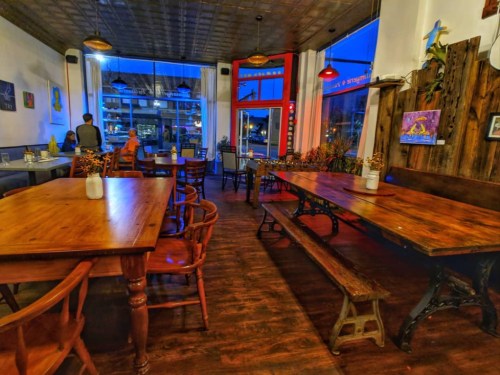 Cozy restaurant interior with wooden tables, chairs, and warm lighting, featuring large windows and a welcoming atmosphere.