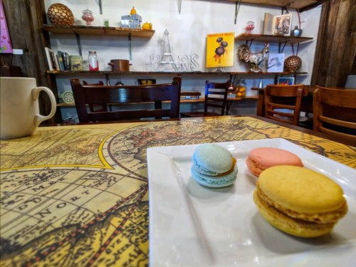 A plate with three colorful macarons sits on a table with a vintage map, surrounded by a cozy café interior.