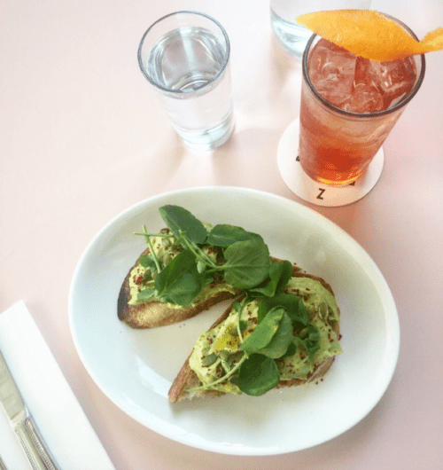 A plate with two avocado toasts topped with greens, a glass of water, and a cocktail with an orange garnish.