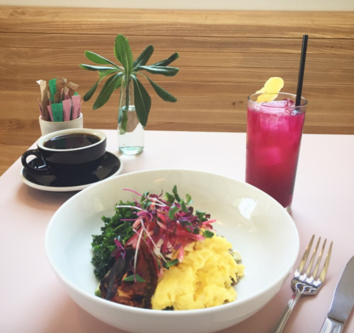 A bowl of scrambled eggs with greens, a coffee cup, a pink drink, and a small plant on a table.