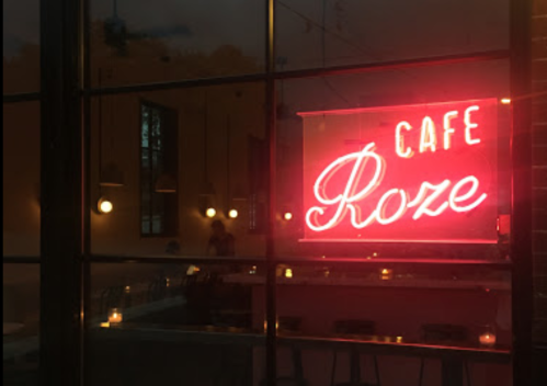 Neon sign reading "Cafe Roze" illuminated in a window, with soft lighting and candles visible inside.