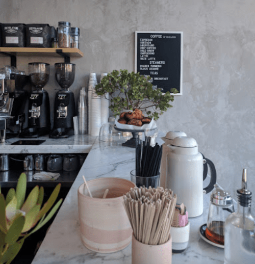 A cozy café counter with coffee machines, pastries, and a plant, featuring a menu board in the background.