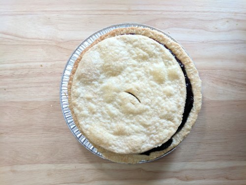 A pie with a golden crust, slightly cracked, sitting in a silver pie tin on a wooden surface.