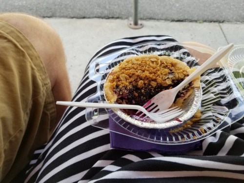 A dessert in a plastic container with a crumb topping, resting on a lap with striped fabric and a leg visible.