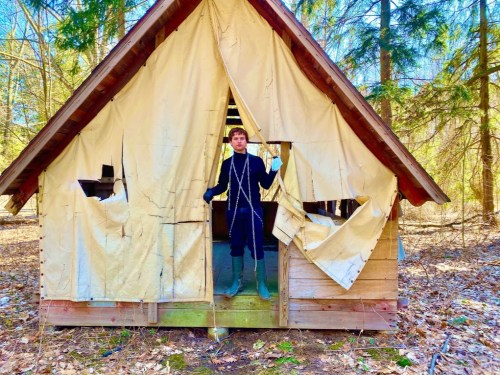 A person stands in the doorway of a rustic cabin with torn fabric, surrounded by trees and fallen leaves.