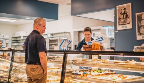 A customer stands at a bakery counter, looking at treats while a staff member reviews an order.