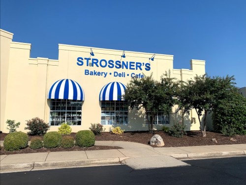 Exterior of Strossner's Bakery, Deli, and Cafe, featuring blue and white striped awnings and landscaped greenery.