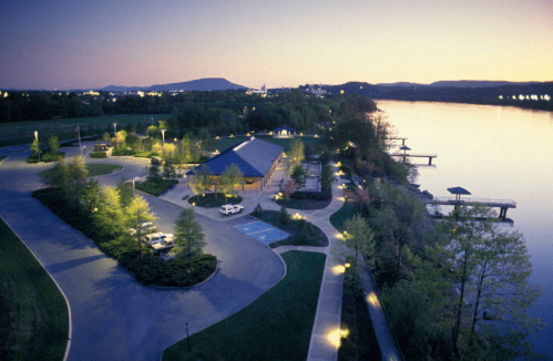 A serene riverside view at dusk, featuring a building, parking area, and illuminated pathways along the water.