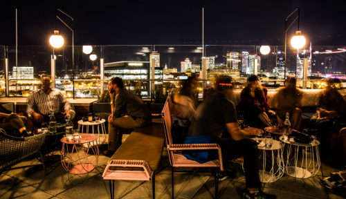 A rooftop bar at night with blurred patrons enjoying drinks and city skyline views in the background.