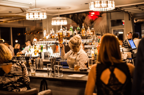 A bartender mixes drinks at a stylish bar, with patrons enjoying their evening in a lively atmosphere.