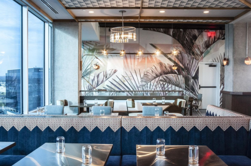 Modern restaurant interior featuring palm leaf wallpaper, stylish lighting, and elegant seating by large windows.