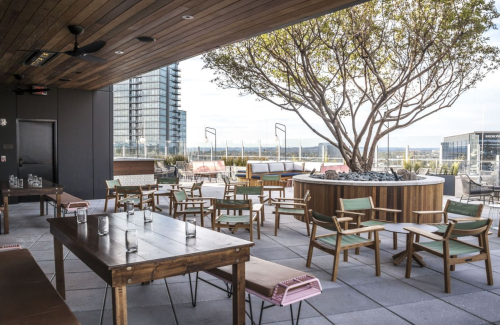 A modern rooftop patio with wooden tables, chairs, and a large tree, overlooking a city skyline and distant hills.