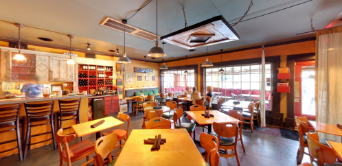 Cozy restaurant interior with wooden tables, chairs, and a bar area, featuring warm lighting and large windows.