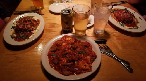 A table with three plates of pasta dishes, two glasses of beer, and a can, all on a wooden surface.