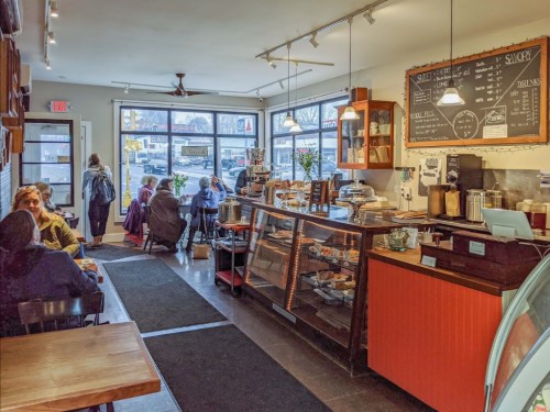 Cozy café interior with a counter displaying pastries, customers seated, and large windows letting in natural light.