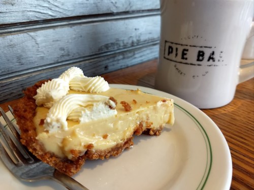 A slice of creamy pie with whipped cream on top, served on a plate next to a coffee mug labeled "PIE BAR."