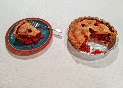 A slice of cherry pie on a plate next to a whole cherry pie in a tin, both on a textured white tablecloth.