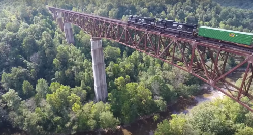 A train crosses a tall, red metal bridge over a river, surrounded by lush green trees.