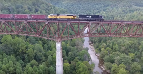 A train crosses a tall, rusted bridge over a river, surrounded by lush green trees and hills.