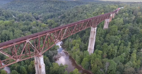 Aerial view of a rusted railway bridge spanning a river, surrounded by dense green trees and hills.