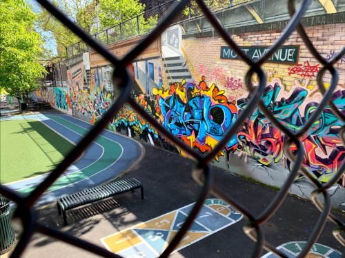 Colorful graffiti covers a wall near a playground, viewed through a chain-link fence.