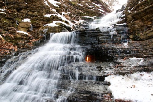 A cascading waterfall flows over rocky terrain, surrounded by snow and ice, with a small cave visible in the rock face.