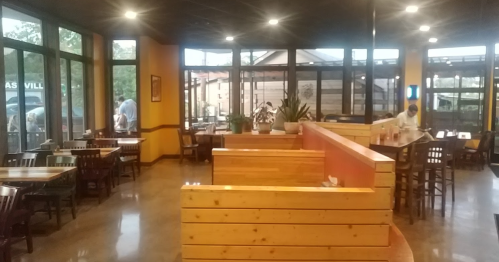 Interior of a restaurant with wooden seating, large windows, and a warm atmosphere. People are visible in the background.