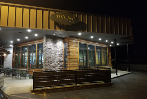 Exterior of Mitchell's restaurant at night, featuring wooden and stone design, illuminated windows, and outdoor seating.