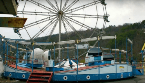 An abandoned amusement park scene featuring a ferris wheel and a derelict boat ride.