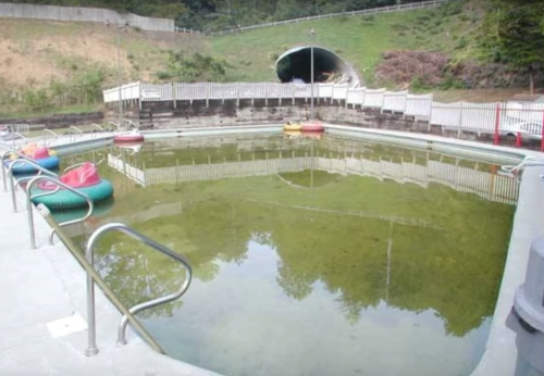 An empty, overgrown pool with green water, surrounded by a concrete deck and a tunnel in the background.