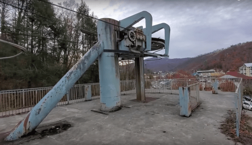 Abandoned ski lift structure with rusted machinery, surrounded by trees and mountains in the background.