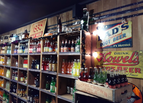 A wooden wall display filled with various colorful soda bottles and vintage signs for root beer and cola.