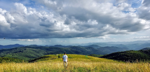 A person stands on a grassy hill, gazing at a vast mountain landscape under a cloudy sky.