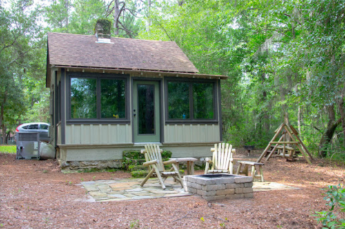 A cozy cabin surrounded by trees, featuring a fire pit and seating area in a natural setting.