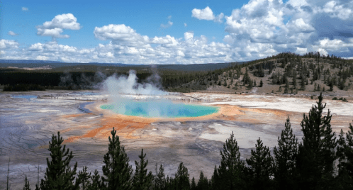 A vibrant geothermal pool surrounded by colorful mineral deposits and lush trees under a blue sky with fluffy clouds.