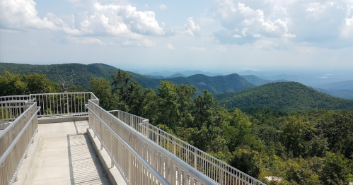 A scenic view from a mountain overlook, featuring lush green hills and a cloudy blue sky in the background.