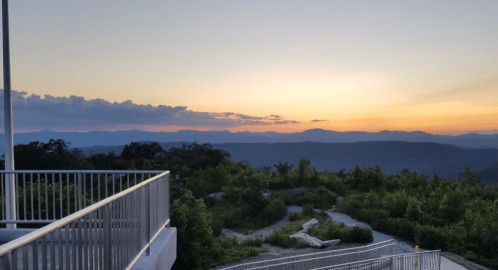 Sunset over mountains with a clear sky, viewed from a scenic overlook surrounded by greenery.