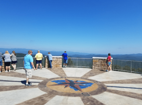 People stand on a scenic overlook with a compass design, gazing at distant mountains under a clear blue sky.