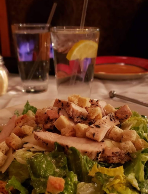 A fresh salad topped with grilled chicken and croutons, with two glasses of water in the background.