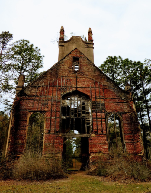 Ruins of a brick church with a tall, crumbling structure surrounded by trees and overgrown vegetation.