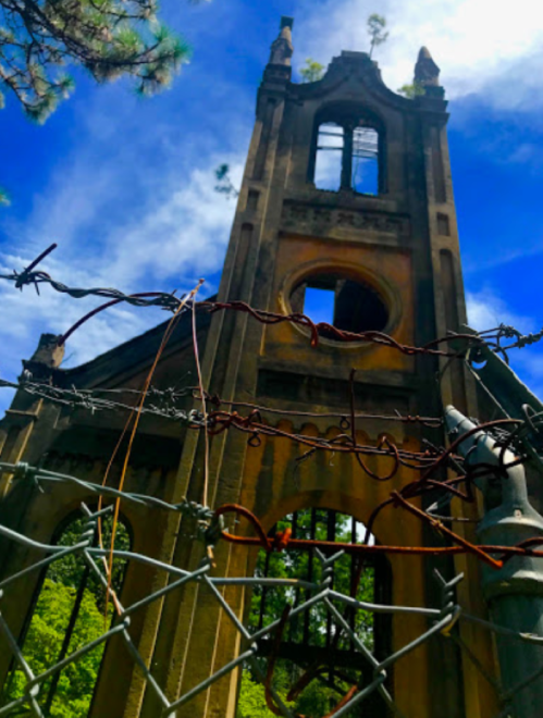A weathered, abandoned building with a tall tower, surrounded by barbed wire and a blue sky with scattered clouds.