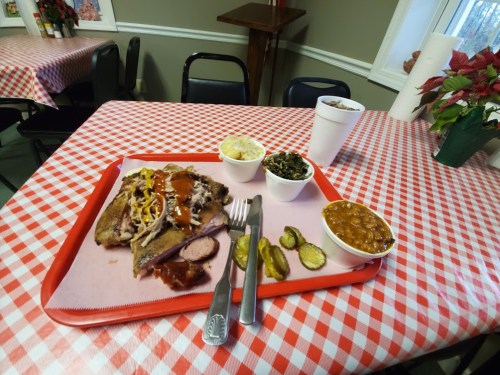 A red-checkered table with a tray of barbecue, sides of coleslaw, greens, baked beans, pickles, and a drink.
